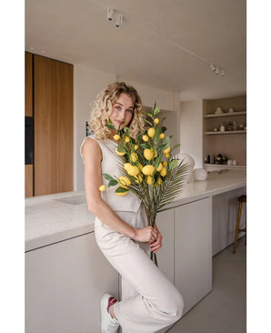 Een vrouw met krullend blond haar, gekleed in een crèmekleurige outfit, leunt tegen een aanrecht en houdt het boeket ‘Kunsttak Lemon 104 cm geel’, versierd met weelderig groen, vast. De moderne keuken is uitgevoerd in neutrale kleuren en heeft houten accenten.