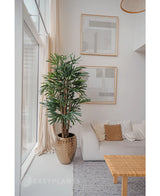 Een Lady Kunstpalm 180 cm is geplaatst bij een groot raam in een lichte, moderne woonkamer met een witte bank, beige kussens, geweven tafel en abstracte kunst. Het voegt een gezellige minimalistische stijl toe met zijn neutrale tinten en groene kunstplant charme.
